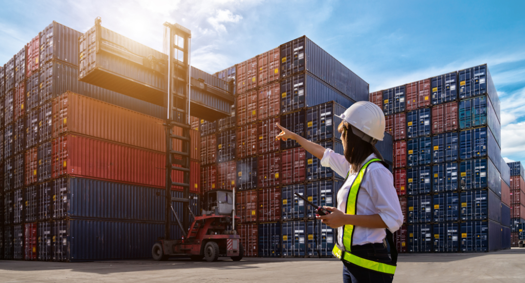 A female worker in a hard hat and safety vest pointing towards stacked shipping containers in a logistics yard under a bright sky